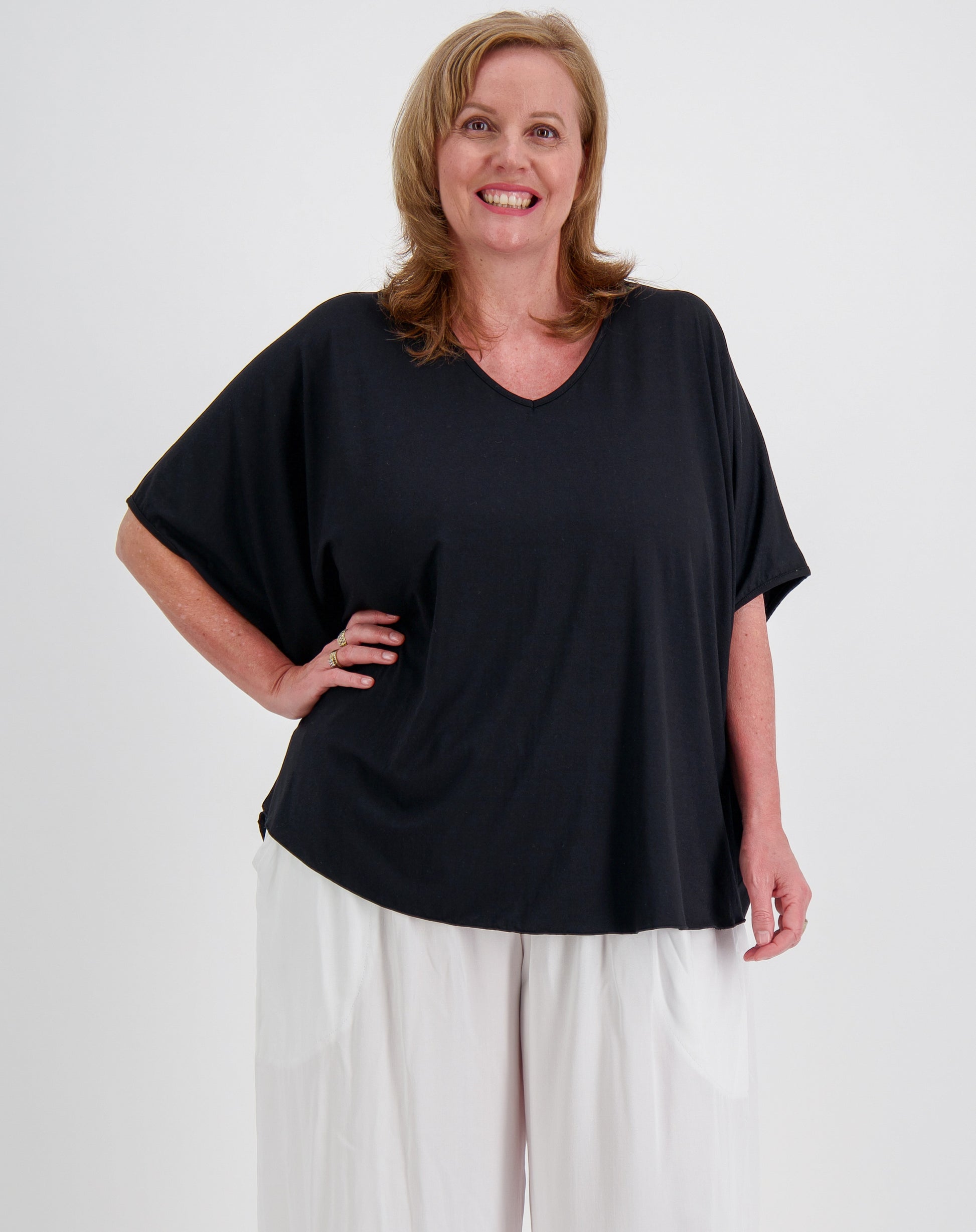 Woman wearing a black curved hem tee, the comfort top by miss seafari and white pants on a plain background