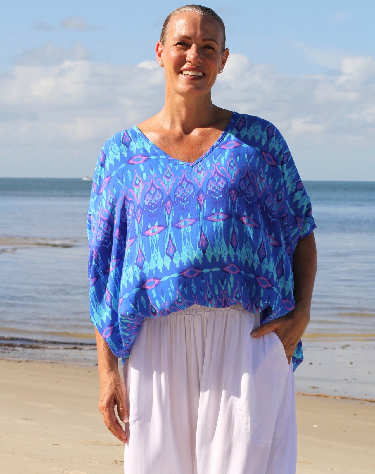 A smiling woman standing on a sandy beach wearing a loose, short-sleeve V-neck top with a blue, purple, and teal ikat pattern, paired with white pants.