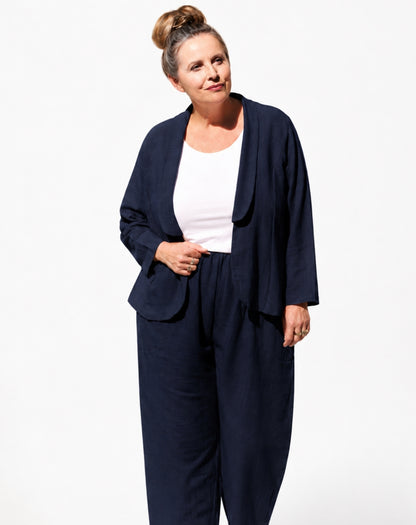 A woman modeling a navy blue two-piece linen look blazer and pant set consisting of a draped open-front jacket and matching relaxed-leg slip on pants, worn over a white tank top, against a plain white studio background.