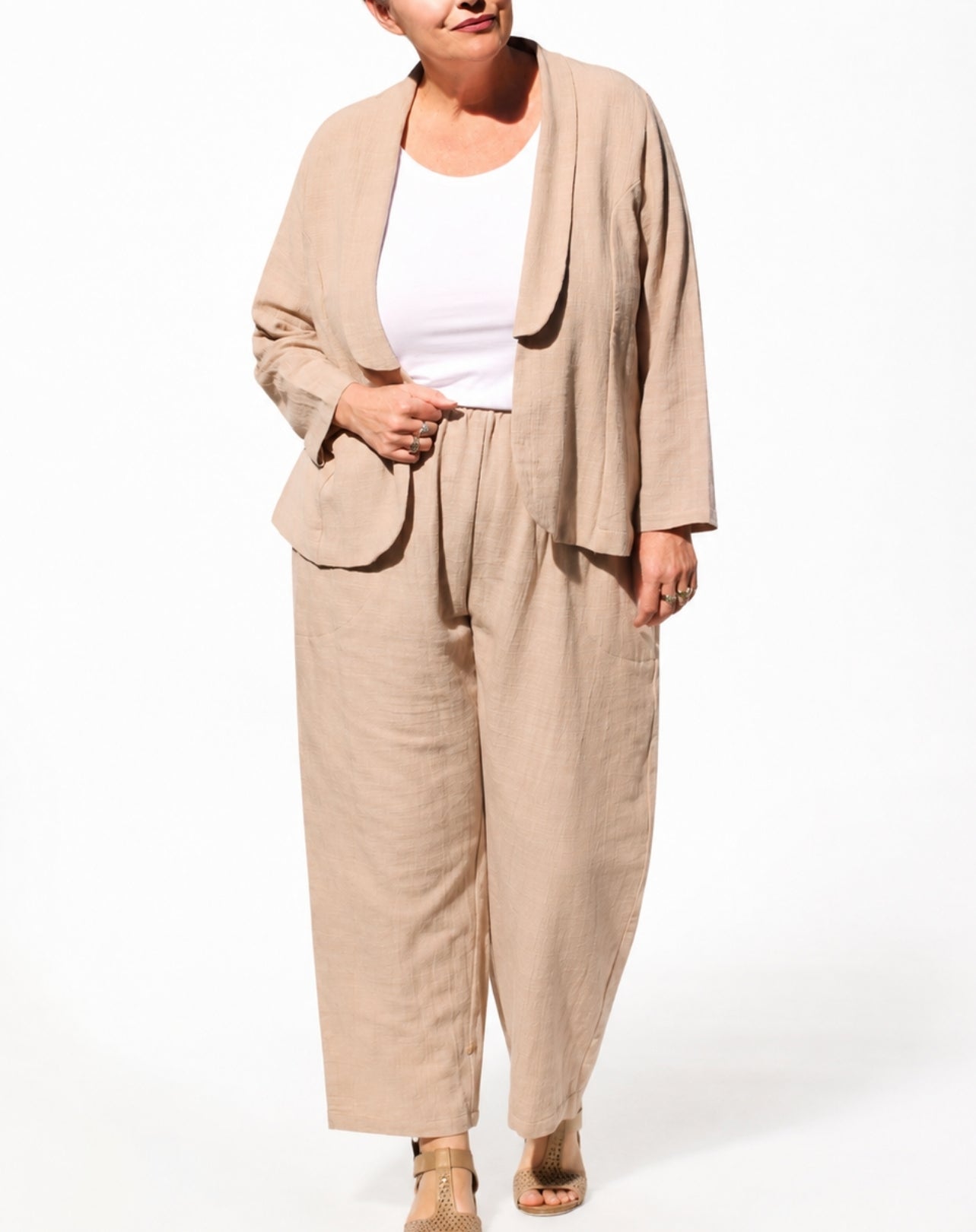A woman modelling a natural  linen two-piece set consisting of a draped open-front jacket and matching wide-leg trousers, styled over a white tank top and tan sandals against a white studio background.