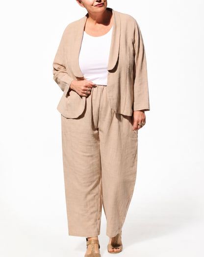 A woman modelling a natural  linen two-piece set consisting of a draped open-front jacket and matching wide-leg trousers, styled over a white tank top and tan sandals against a white studio background.