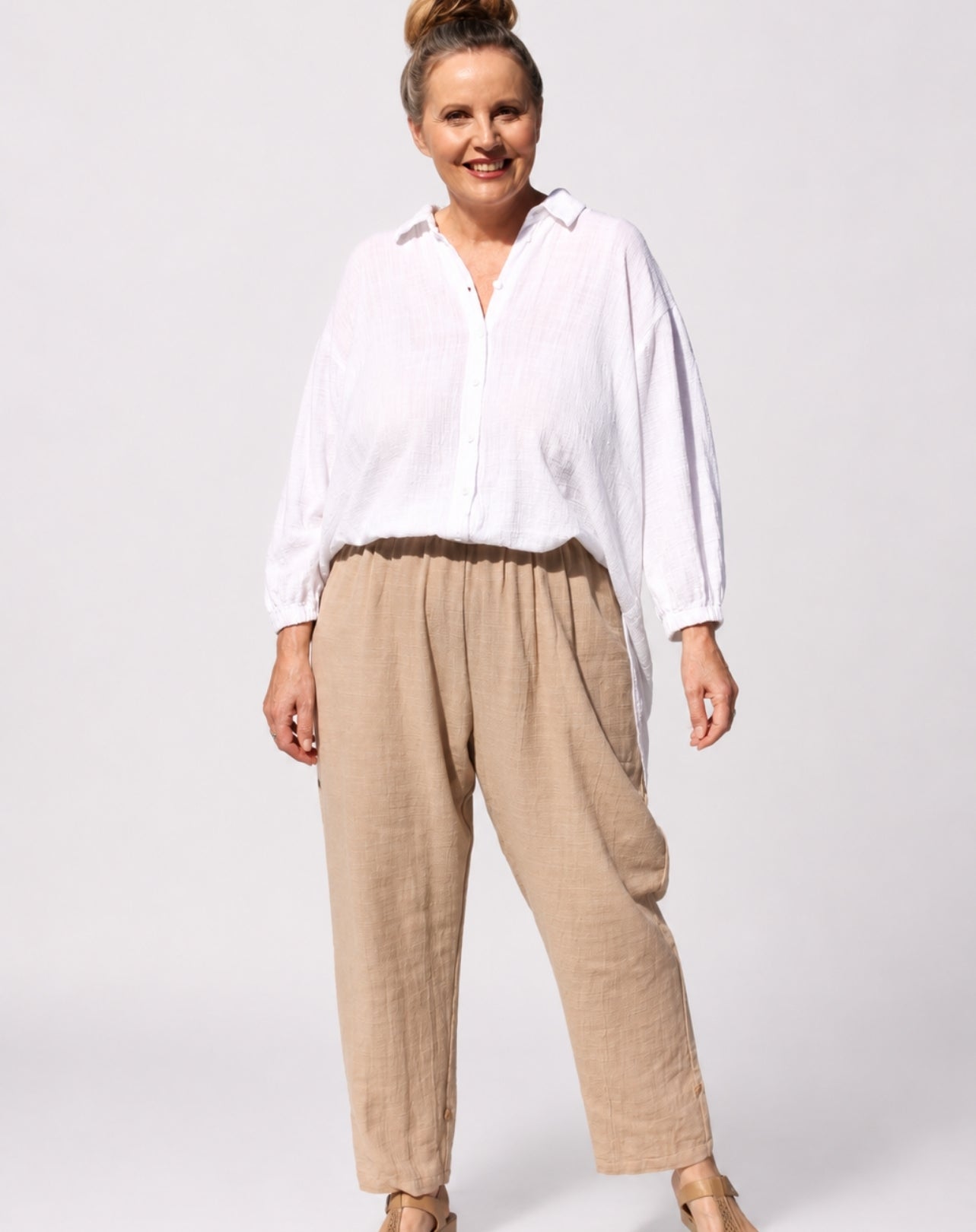 A woman modelling a white long-sleeve button-up shirt and beige natural linen trousers against a light grey studio background.