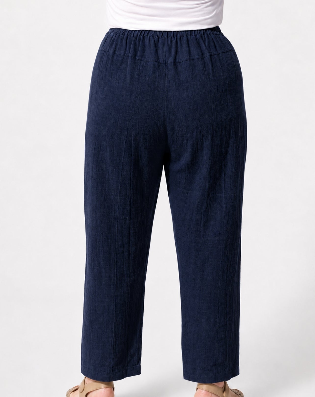 A person wearing dark navy relaxed straight-leg cropped pants with an elastic waistband, paired with a white top and beige sandals, shown from the waist down on a light grey background.