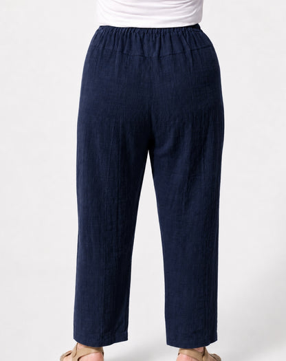 A person wearing dark navy relaxed straight-leg cropped pants with an elastic waistband, paired with a white top and beige sandals, shown from the waist down on a light grey background.