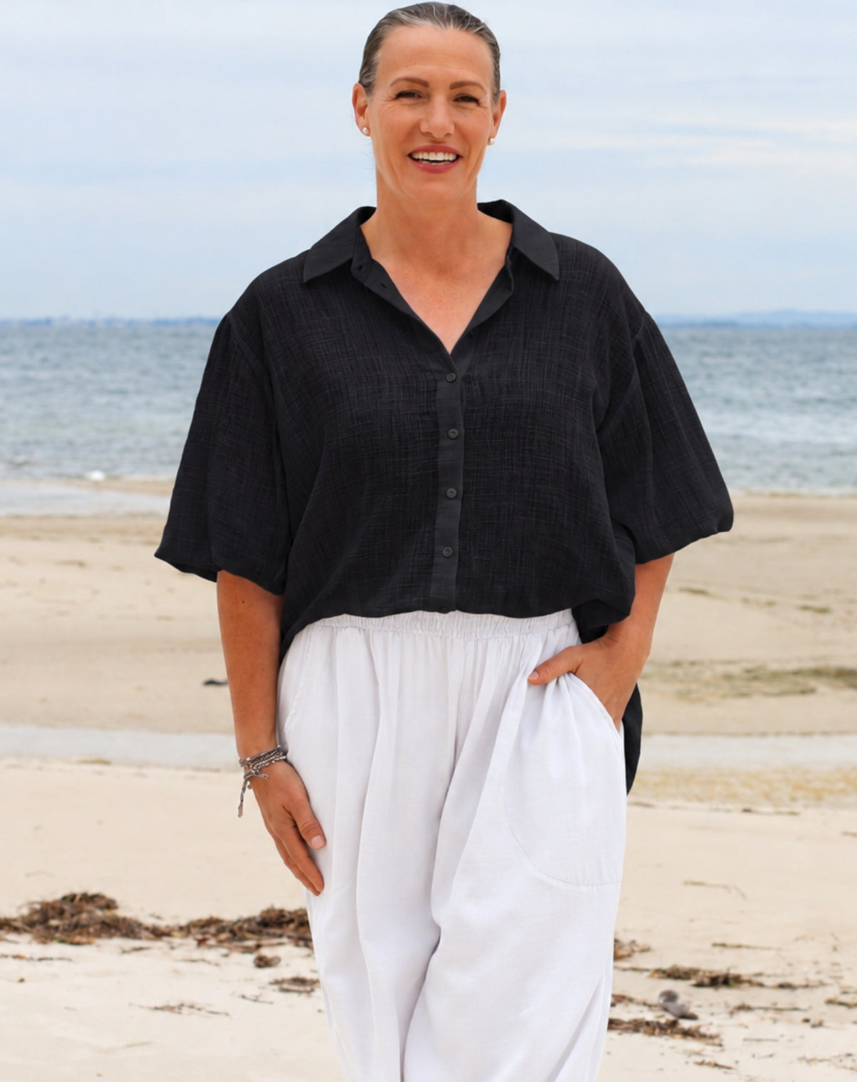 A woman standing on a sandy beach wearing a loose black short-sleeve button-up shirt and white wide-leg pants, with the ocean and pale sky in the background.
