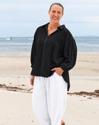 A woman standing on a sandy beach wearing a loose black button-up shirt and white wide-leg pants, with the ocean and sky in the background.