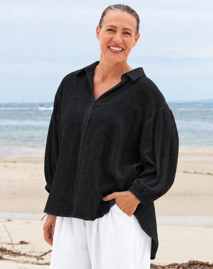 A smiling woman standing on a sandy beach wearing a loose black long-sleeve button-up shirt and white pants, with the ocean and cloudy sky in the background.
