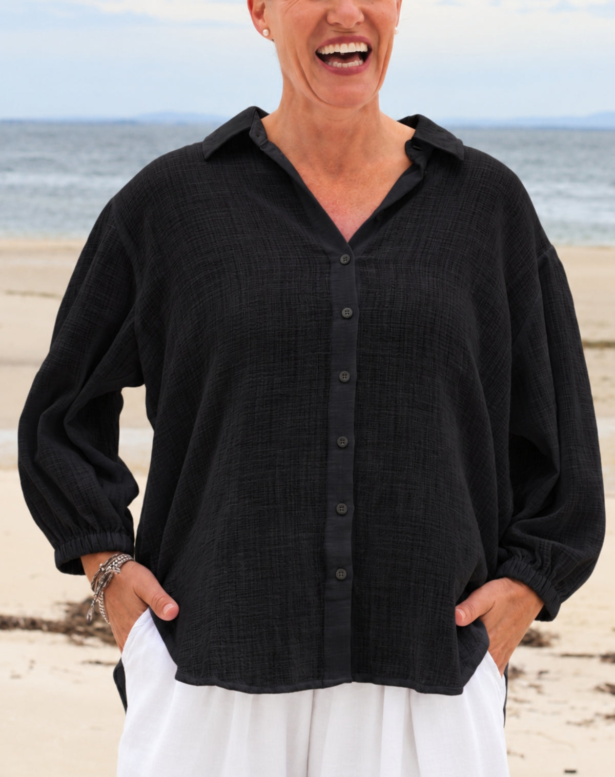 A woman wearing a black textured long-sleeve button-up shirt and white pants, standing on a sandy beach with the ocean in the background.