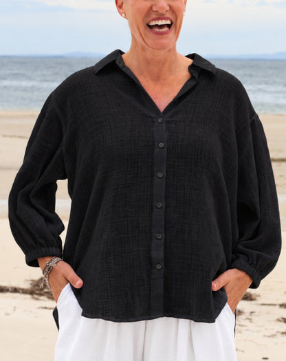 A woman wearing a black textured long-sleeve button-up shirt and white pants, standing on a sandy beach with the ocean in the background.