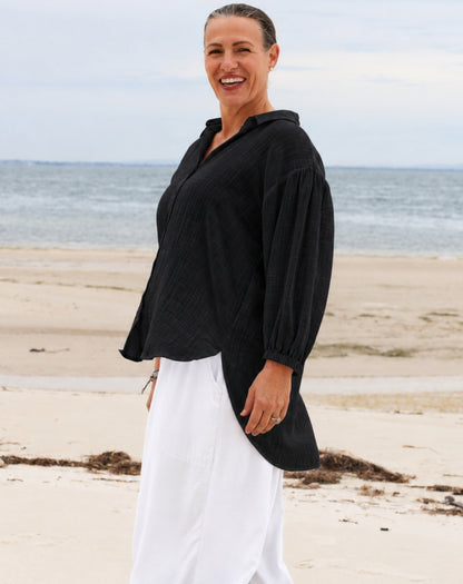 A woman standing on a sandy beach wearing a loose cotton black long-sleeve top and white wide-leg pants, with the ocean and cloudy sky in the background.