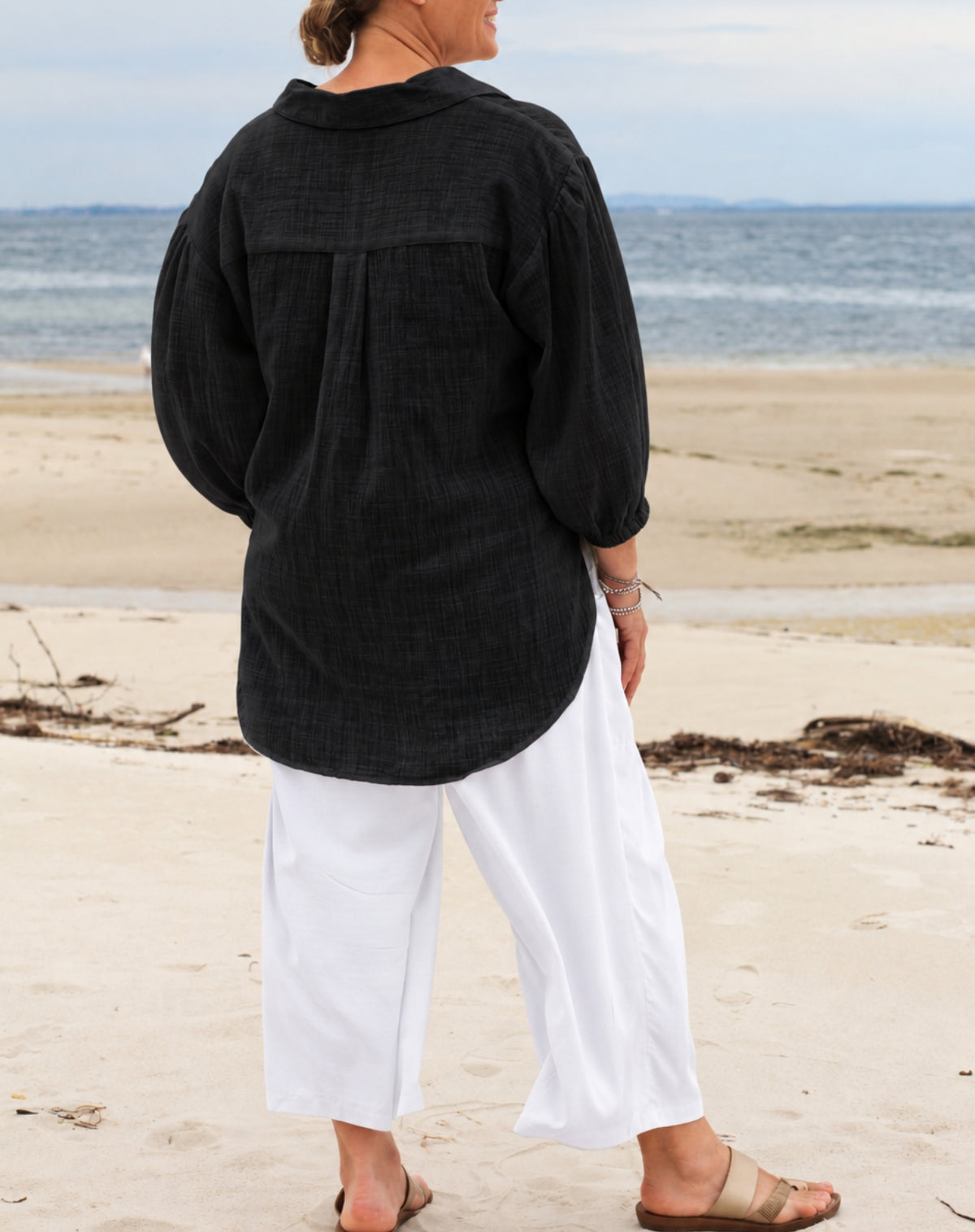 A woman standing on a sandy beach with the ocean in the background, wearing a loose black long-sleeve top and white cropped pants.