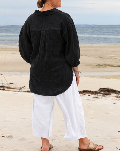 A woman standing on a sandy beach with the ocean in the background, wearing a loose black long-sleeve top and white cropped pants.