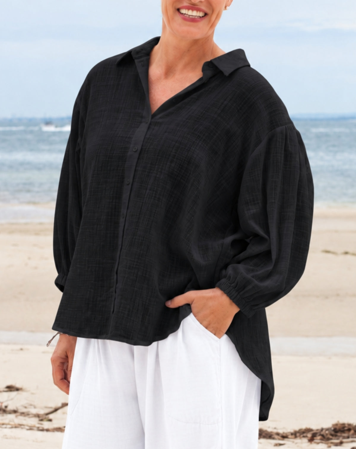 A woman wearing a black cotton textured long-sleeve button-up shirt and white pants, standing on a sandy beach with the ocean in the background.
