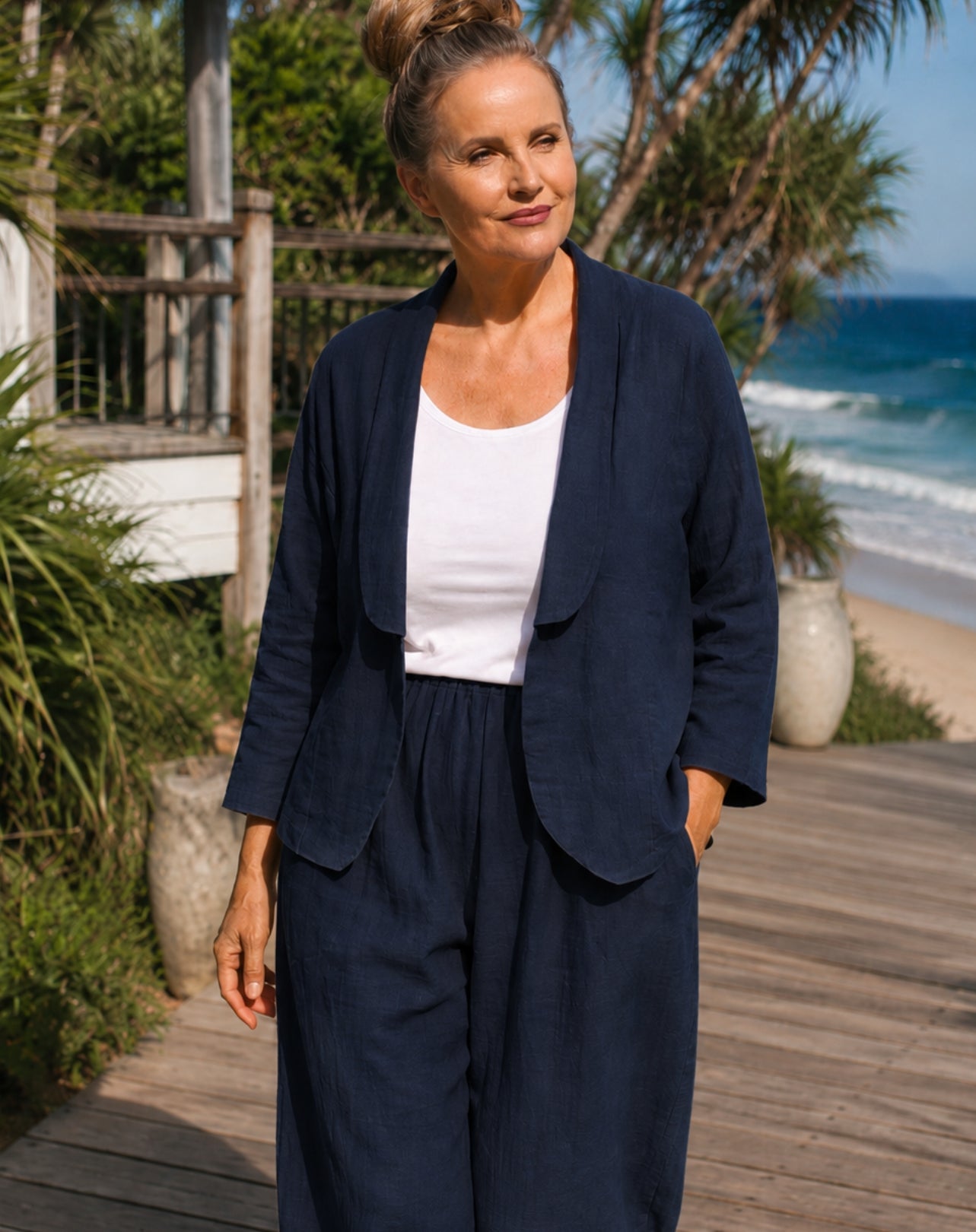 A woman wearing a navy blue linen-blend two-piece set consisting of a draped open-front blazer jacket and matching wide-leg pants, styled with a white scoop tank top, standing on a wooden boardwalk with a beach and ocean background.