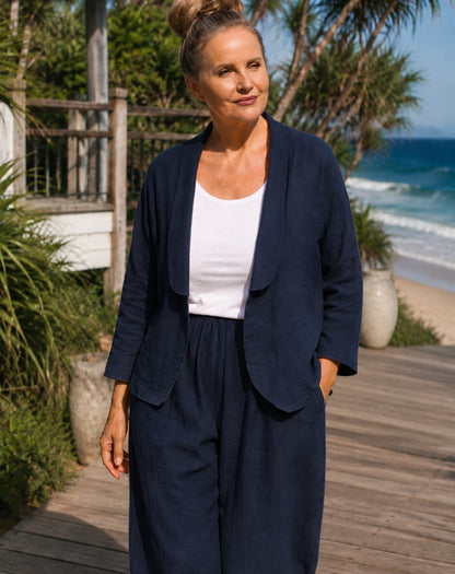 A woman wearing a navy blue linen-blend two-piece set consisting of a draped open-front blazer jacket and matching wide-leg pants, styled with a white scoop tank top, standing on a wooden boardwalk with a beach and ocean background.