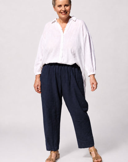 A woman modelling a white long-sleeve button-up shirt and dark navy straight leg trousers, standing against a light grey studio background.