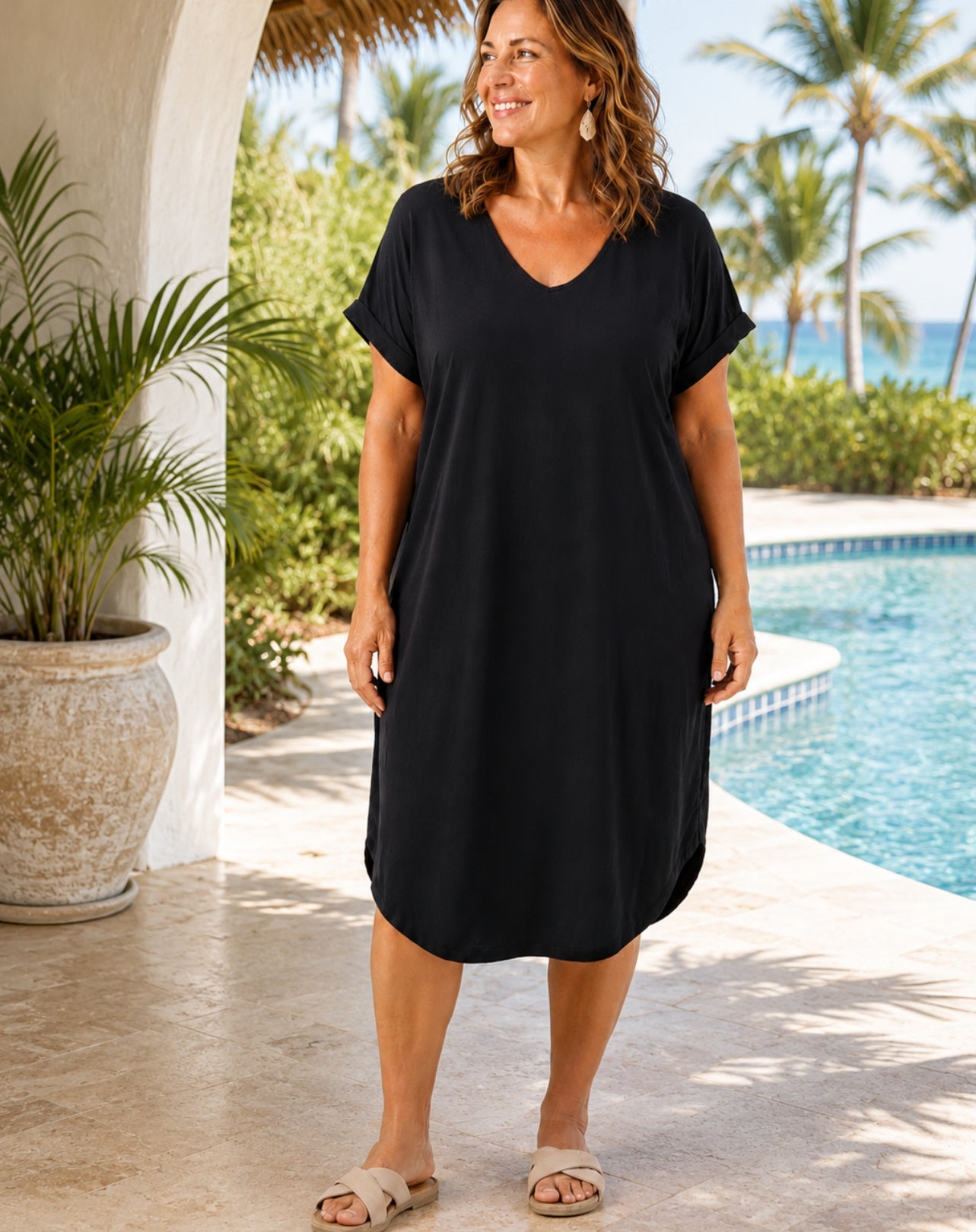 A woman wearing a loose black short-sleeve V-neck dress stands on a poolside patio with palm trees and a thatched roof in the background.
