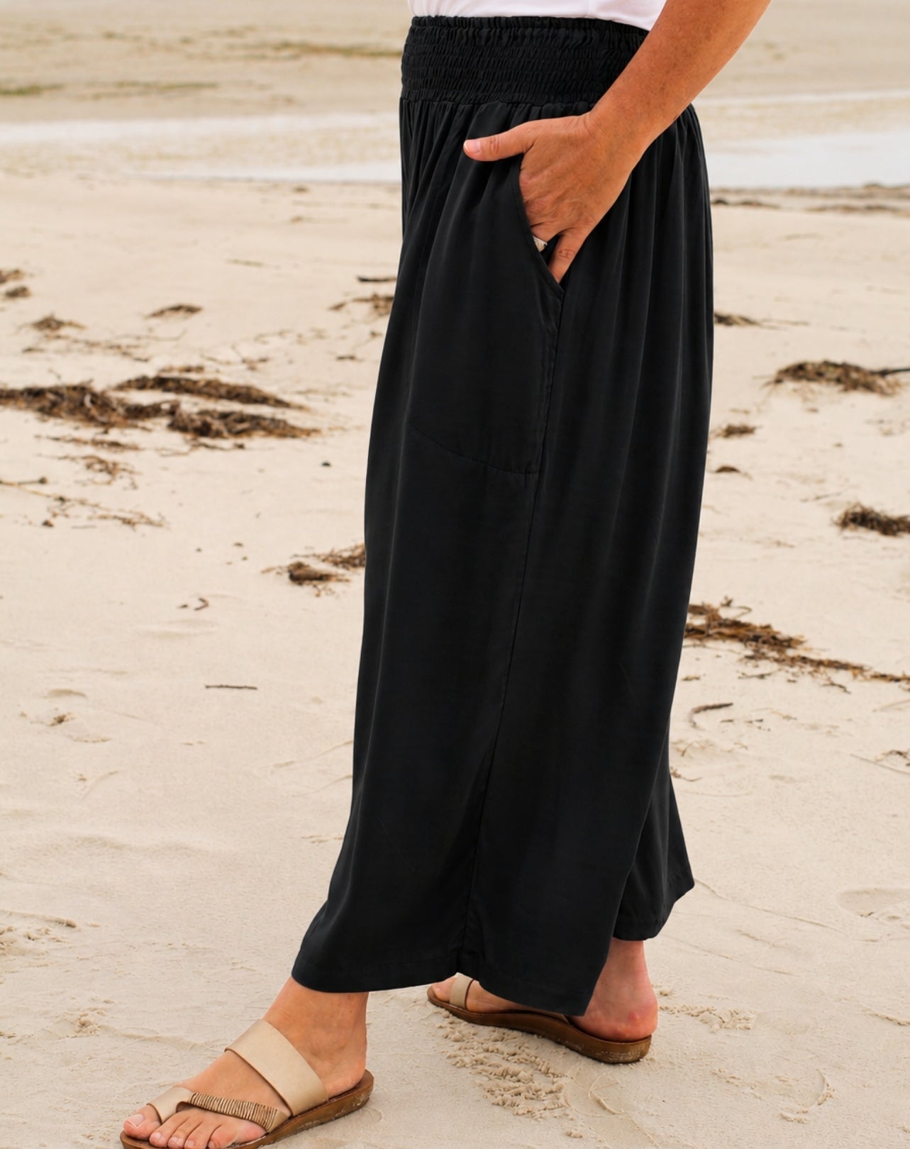 A person wearing a black wide leg crop pant with side pockets, standing on a sandy beach.