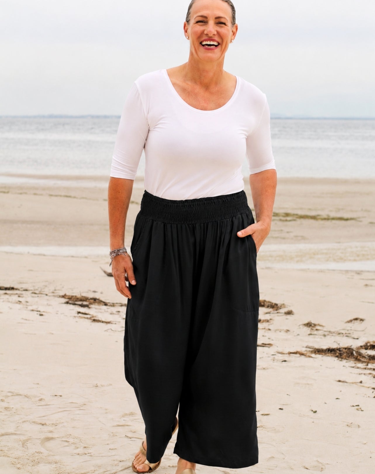 A woman standing on a sandy beach wearing a white short-sleeve scoop-neck top and black wide-leg pants with side pockets.