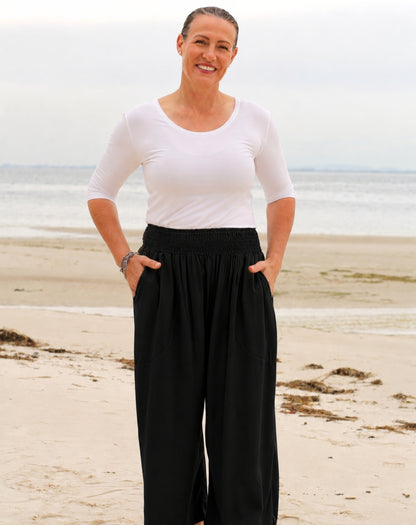 A woman standing on a sandy beach wearing a white short-sleeve scoop-neck top and black wide-leg pants with side pockets.