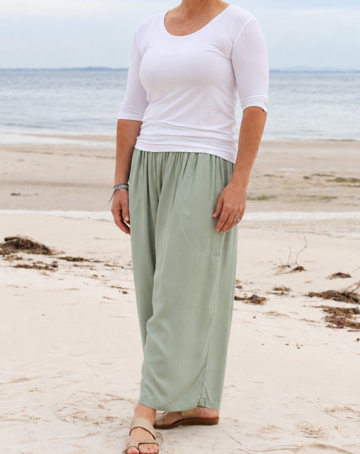 A woman standing on a sandy beach wearing a white three-quarter-sleeve scoop-neck top and light mint green wide-leg pants, paired with tan sandals.