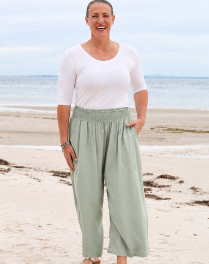 A woman standing on a sandy beach wearing a white short-sleeve scoop-neck top and light sage green wide-leg pants with an elastic waistband.