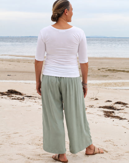 A woman standing on a sandy beach with the ocean in the background, wearing a fitted white short-sleeve top and loose sage green wide-leg pants.