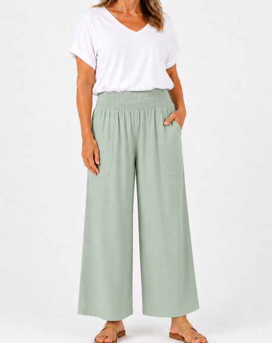 A woman modelling a pair of mint green wide-leg cropped pants with an elastic waistband and side pockets, paired with a white V-neck t-shirt and flat sandals, against a plain white background.
