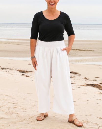 A woman standing on a sandy beach wearing a black short-sleeve top and white wide-leg pants, with her hands in the pockets of the pants.