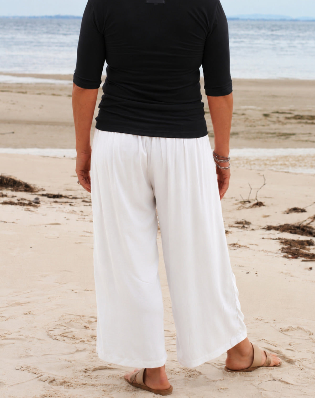 A person standing on a sandy beach facing away from the camera, wearing loose white wide-leg pants and a black short-sleeve top, with the ocean in the background.