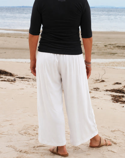 A person standing on a sandy beach facing away from the camera, wearing loose white wide-leg pants and a black short-sleeve top, with the ocean in the background.
