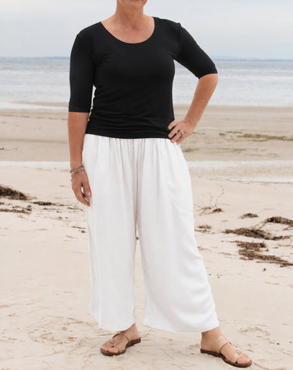 A woman standing on a sandy beach wearing a black three-quarter-sleeve scoop-neck top and white wide-leg pants, paired with brown sandals.