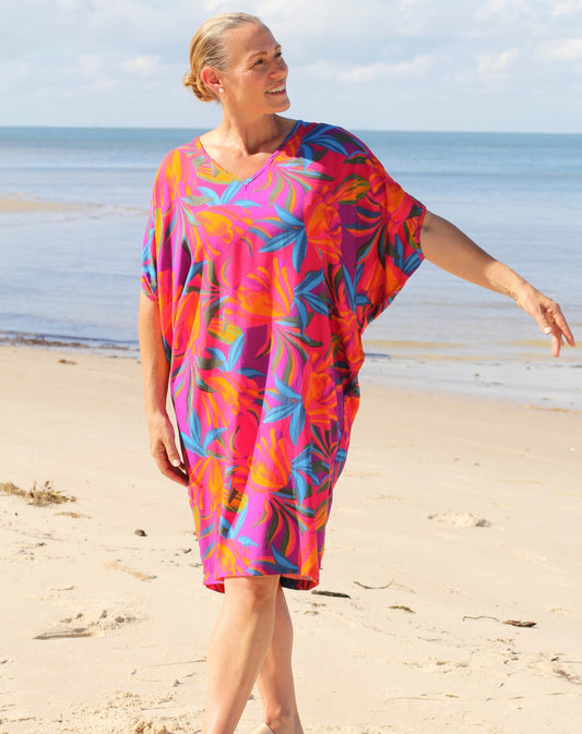 A woman wearing a vibrant, multicolored tropical leaf print kaftan dress walks on a sandy beach with the ocean and sky in the background.