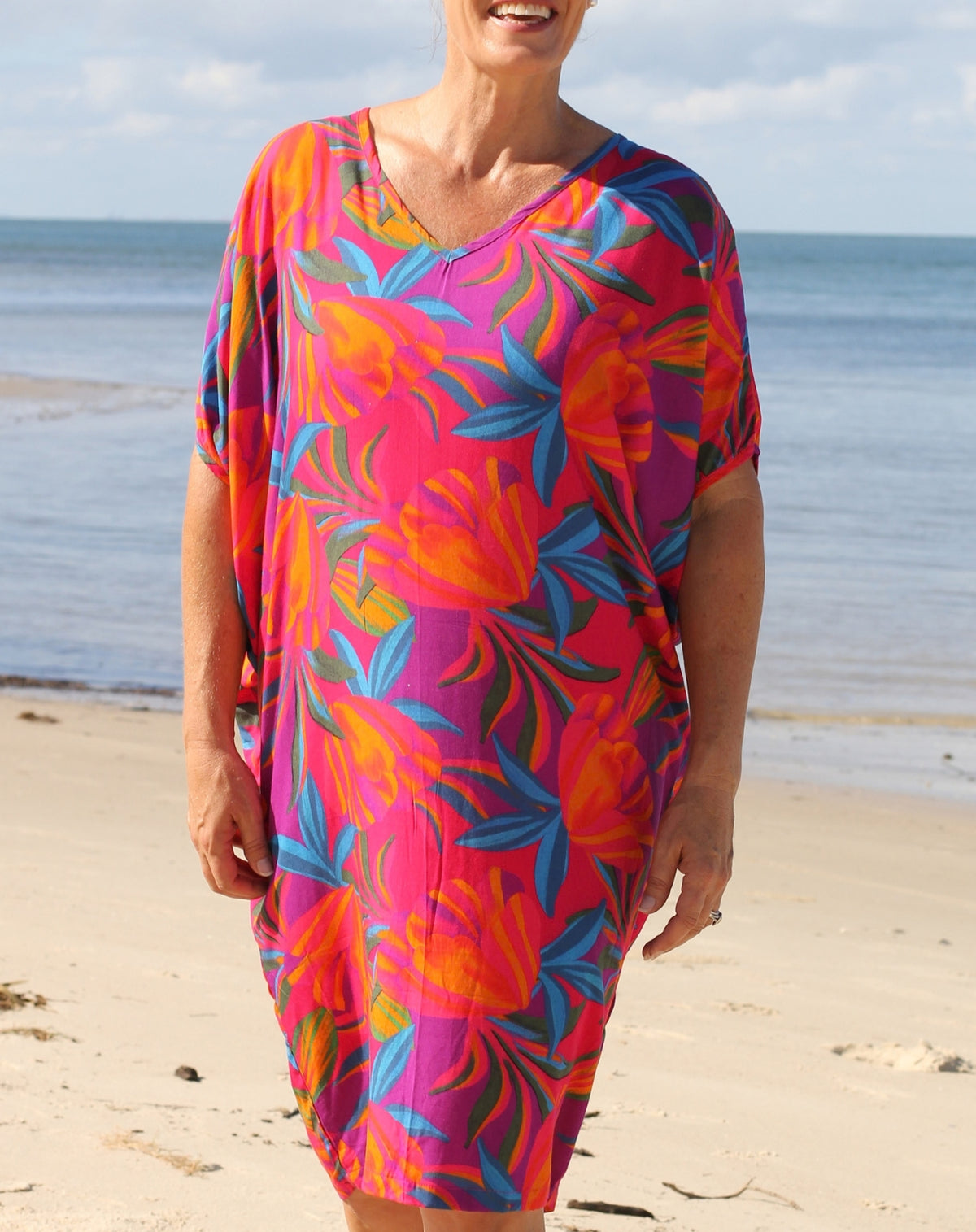 A woman wearing a vibrant tropical floral print kaftan dress stands on a sandy beach with the ocean and sky in the background. The dress features a bold pattern of pink, orange, purple, and blue leaves and flowers.