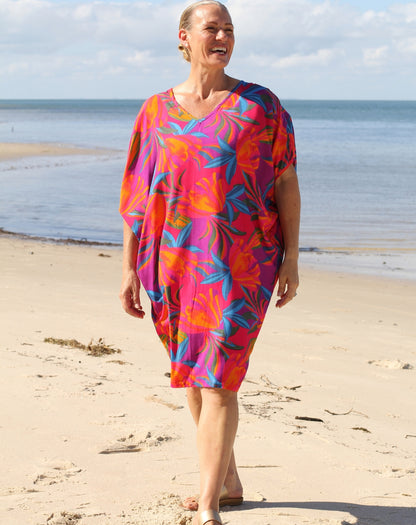 A woman walking on a sandy beach wearing a loose, short kaftan dress with a vibrant tropical floral pattern in pink, orange, blue, and purple.