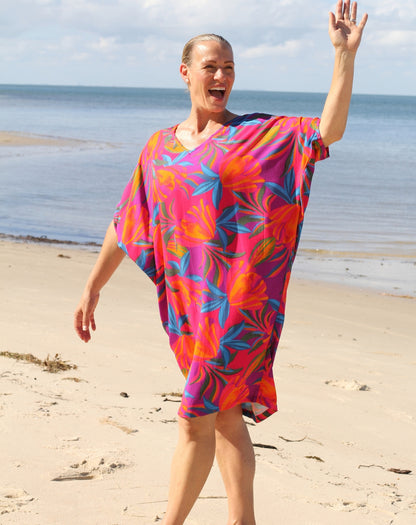 A woman smiling and waving on a sandy beach while wearing a vibrant, short kaftan-style cover-up dress with a bold floral pattern in pink, orange, and blue.