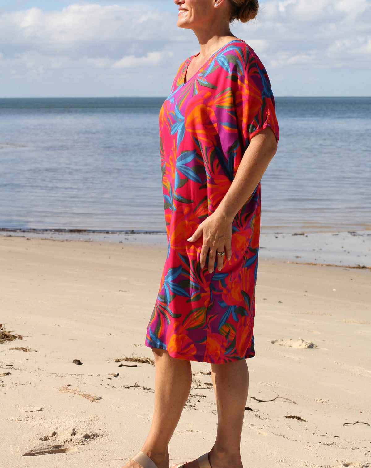 A woman wearing a vibrant, short-sleeved V-neck dress with a bold tropical floral pattern in red, orange, pink, and blue, standing on a sandy beach with the ocean and sky in the background.