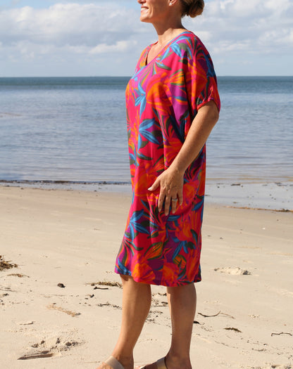 A woman wearing a vibrant, short-sleeved V-neck dress with a bold tropical floral pattern in red, orange, pink, and blue, standing on a sandy beach with the ocean and sky in the background.