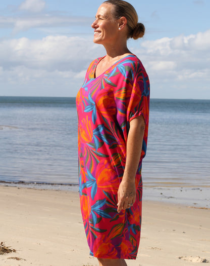 A woman wearing a vibrant, multicolored tropical floral print kaftan dress stands on a sandy beach with the ocean and cloudy sky in the background.