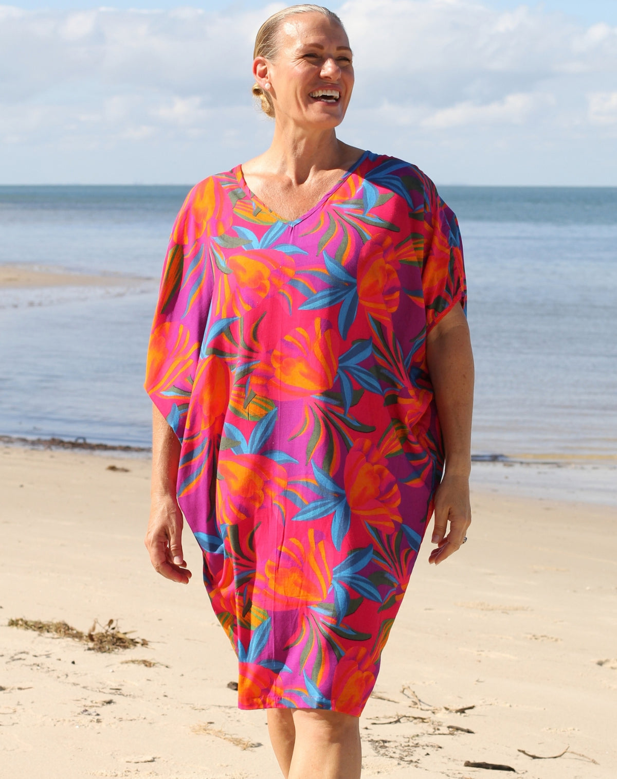 A woman wearing a vibrant tropical floral print kaftan dress walks on a sandy beach with the ocean and cloudy sky in the background.