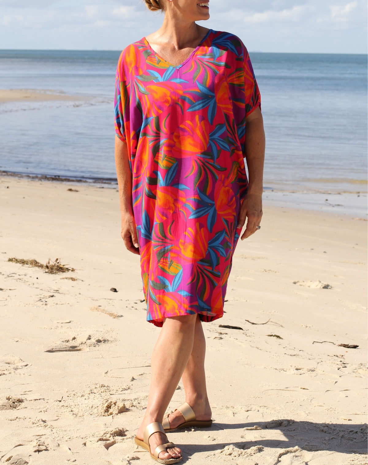 A woman wearing a vibrant, multicolored tropical print kaftan dress stands on a sandy beach with the ocean in the background.