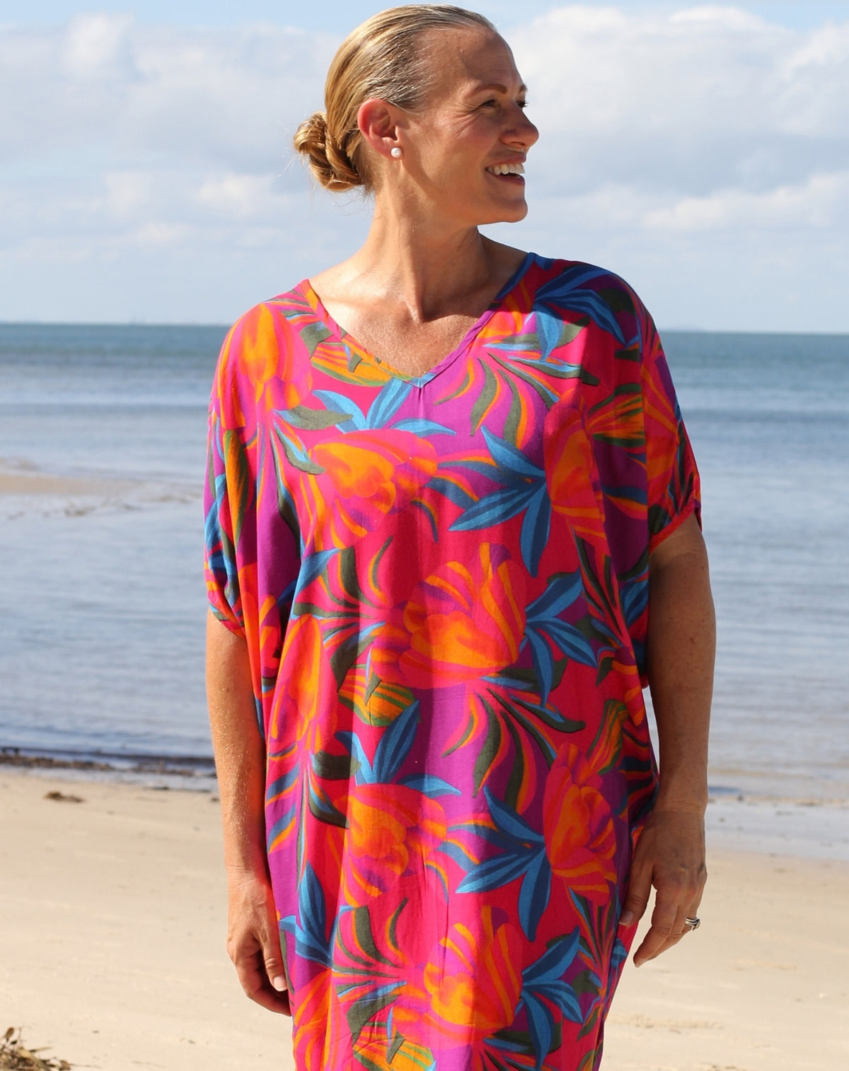 A woman wearing a vibrant tropical print kaftan dress stands on a sandy beach with the ocean and cloudy sky in the background.