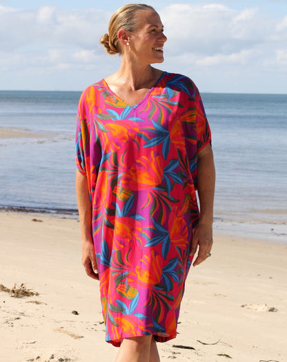 A woman wearing a vibrant tropical floral print kaftan dress stands on a sandy beach with the ocean and cloudy sky in the background.