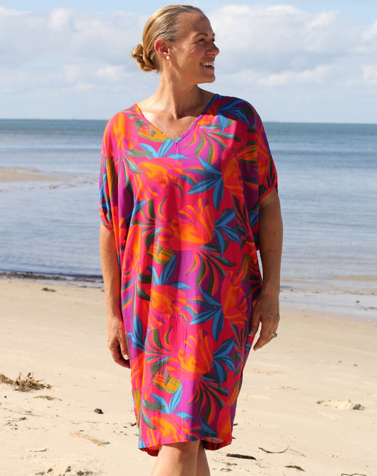 A woman wearing a vibrant tropical floral print kaftan dress stands on a sandy beach with the ocean and cloudy sky in the background.