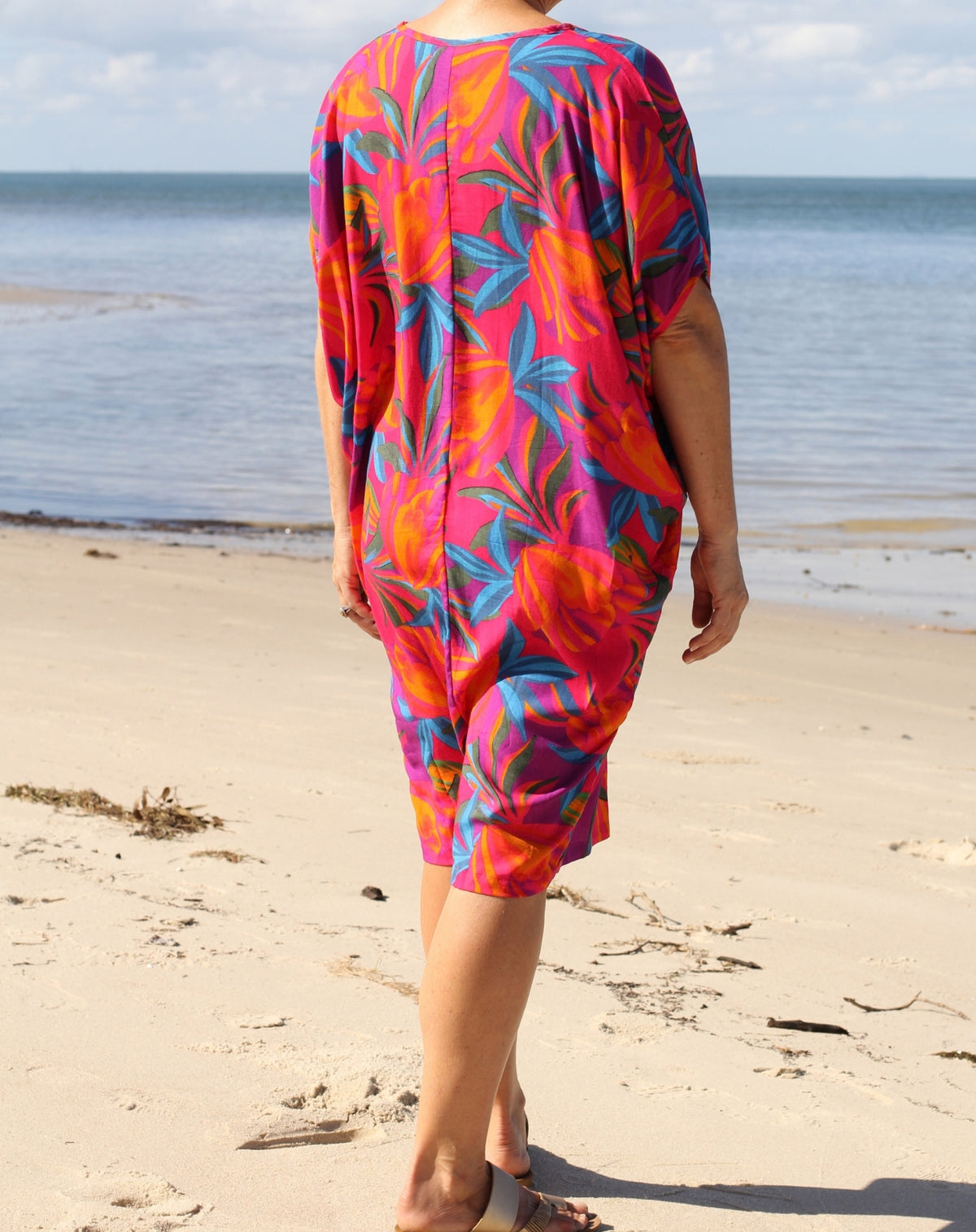 A woman viewed from behind wearing a vibrant tropical floral print cover-up dress with pink, orange, and teal colors, standing on a sandy beach with the ocean in the background.