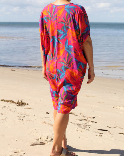 A woman viewed from behind wearing a vibrant tropical floral print cover-up dress with pink, orange, and teal colors, standing on a sandy beach with the ocean in the background.