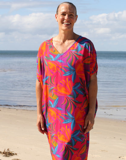 A smiling woman stands on a sandy beach wearing a loose, short-sleeve V-neck dress with a bold tropical floral print in pink, orange, blue, and purple.