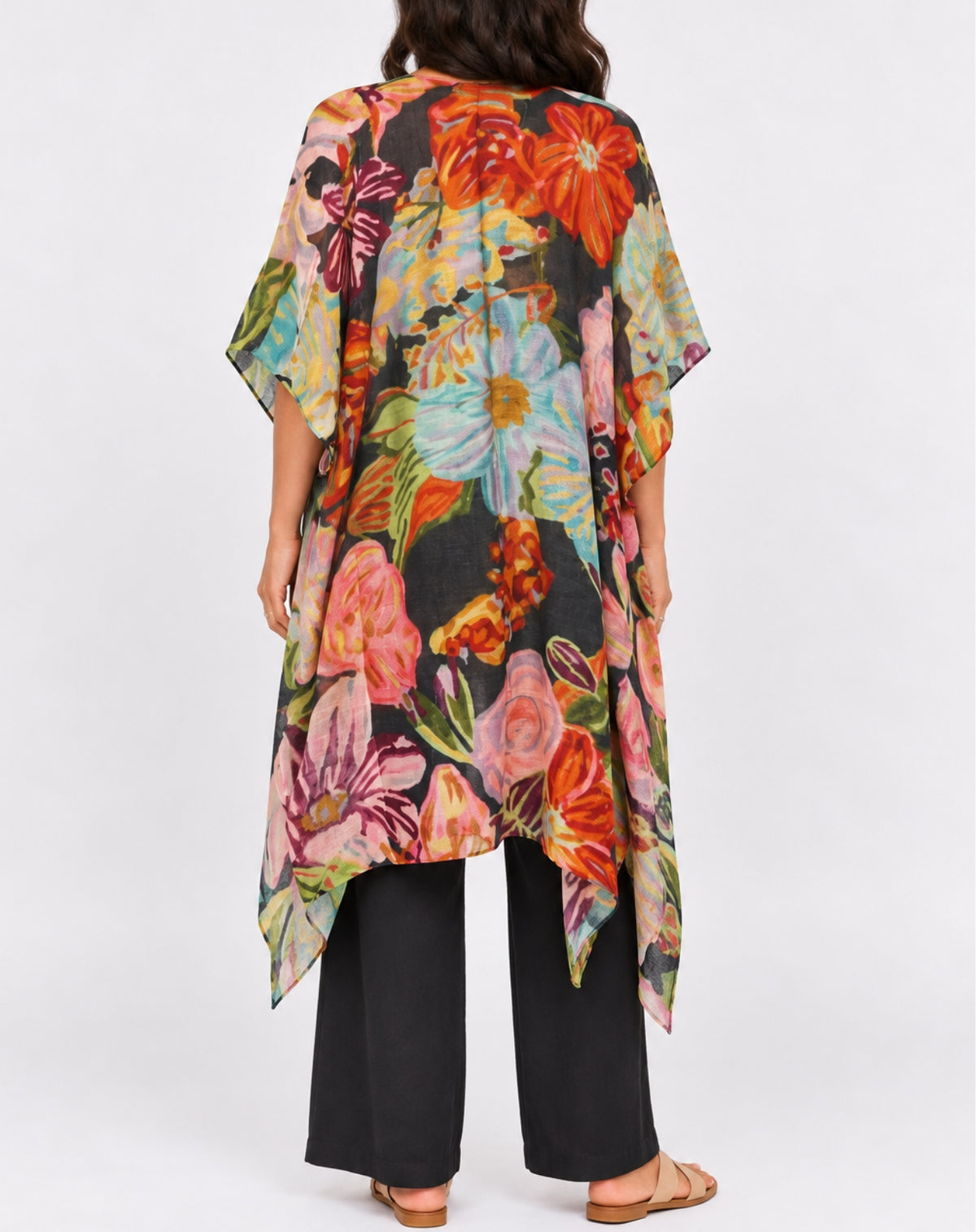 A woman viewed from the back wearing a loose, short-sleeve floral kimono-style top with a black base and large multicolored flowers, paired with black wide-leg pants and tan sandals, against a light gray studio background.