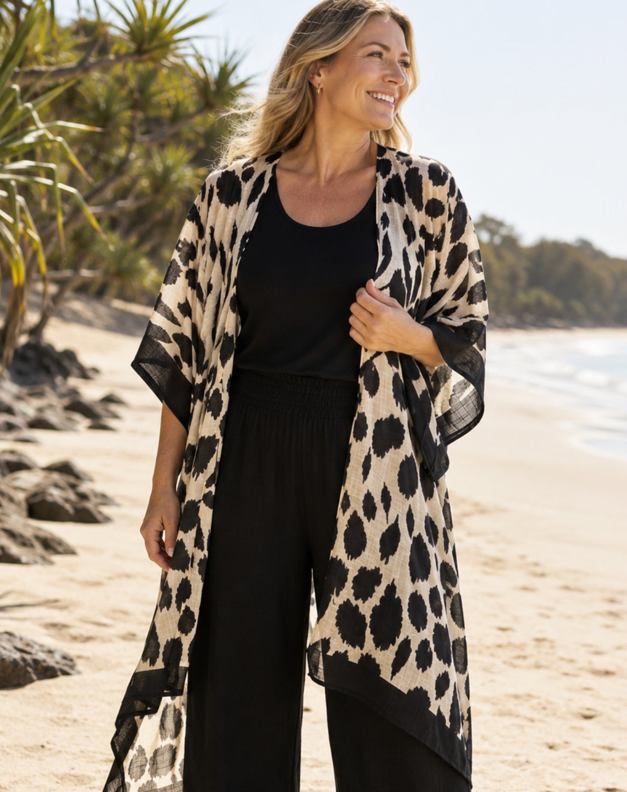 A woman standing on a sandy beach wearing a black and beige animal print open-front kimono over a black resort outfit.