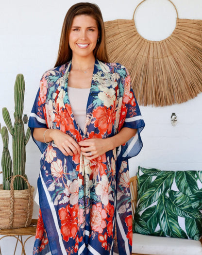 Woman wearing a blue hibiscus floral kimono in a room with decorative plants and wall art.